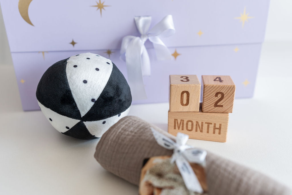 Black and white patterned ball, wooden blocks with '0-2 Month', and a baby blanket on a light surface.